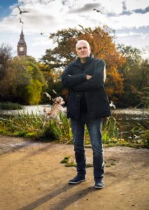 Man standing with arms crossed with a pad and dog in the background and beyond that Big Ben.